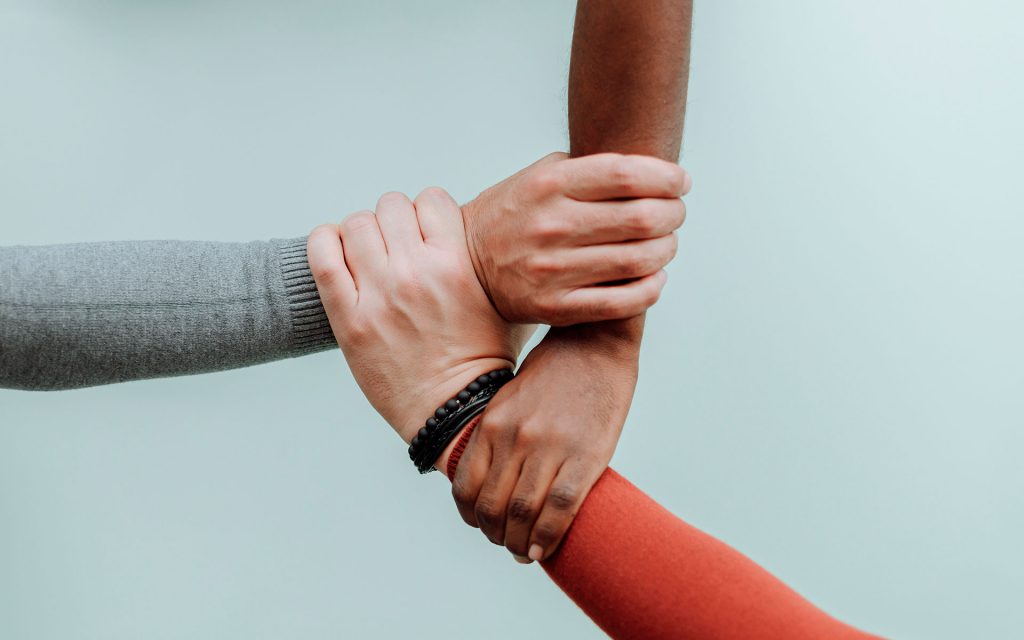 Braços de três pessoas, de etnias aparentemente diferentes, unidos pelas mãos.